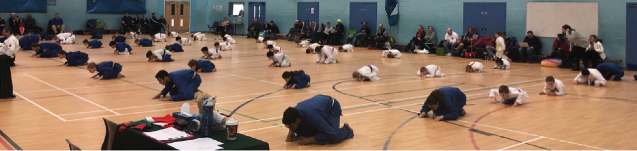 A large group of students in gi kneeling and bowing on a sports-hall floor