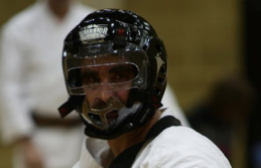 A student in a black headguard with a clear face cage, in white gi